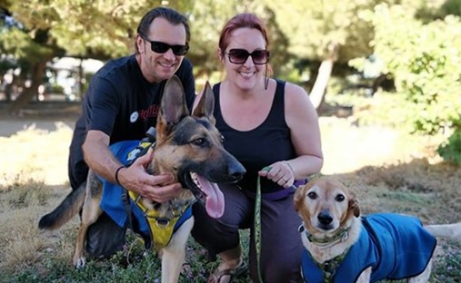 A man and a woman kneel down, in front of them are two dogs